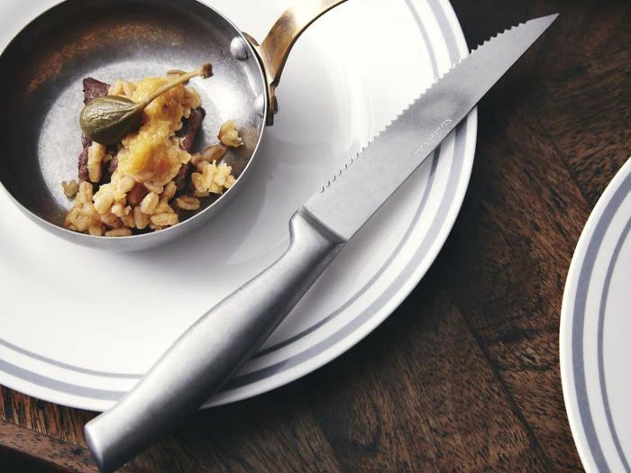 En lille pande med ris, kød og en kapers ligger på en tallerken ved siden af en savtakket steakkniv på et træbord.