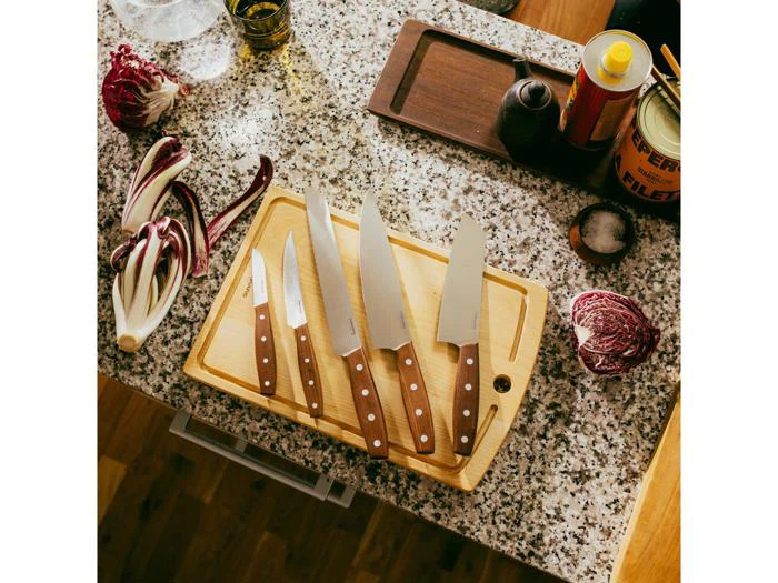 Et sæt med fem køkkenknive med træhåndtag på et skærebræt, omgivet af radicchio, krydderier og madolie på en bordplade af granit.