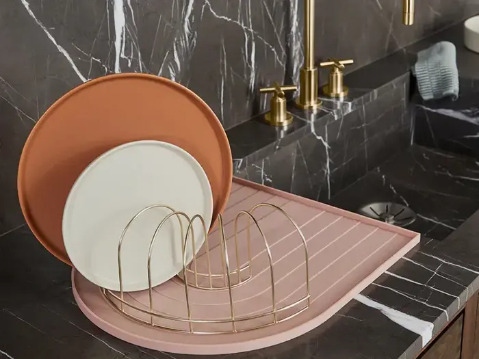 Et lyserødt opvaskestativ med to tallerkener, en hvid og en brun, står på en køkkenbordplade i sort marmor ved siden af en vandhane i guld.