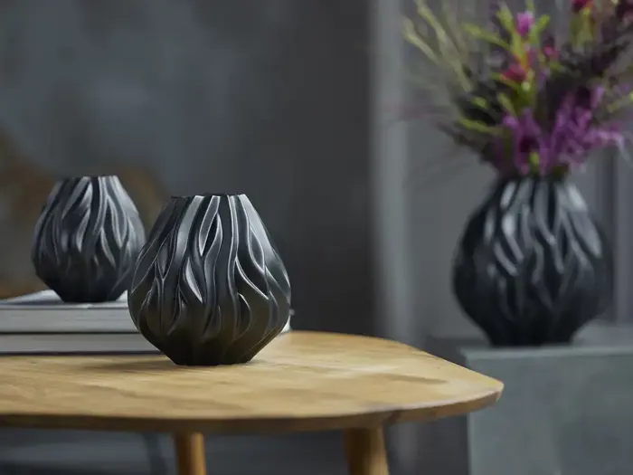 En sort, skulpturel vase med et bølget flammelignende design står på et træbord; en anden vase med blomster er sløret i baggrunden.