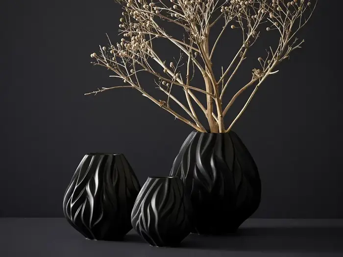 Tre sorte vaser med et bølget tekstureret design står på en mørk overflade; en vase indeholder tørre, beige grene mod en mørk baggrund.