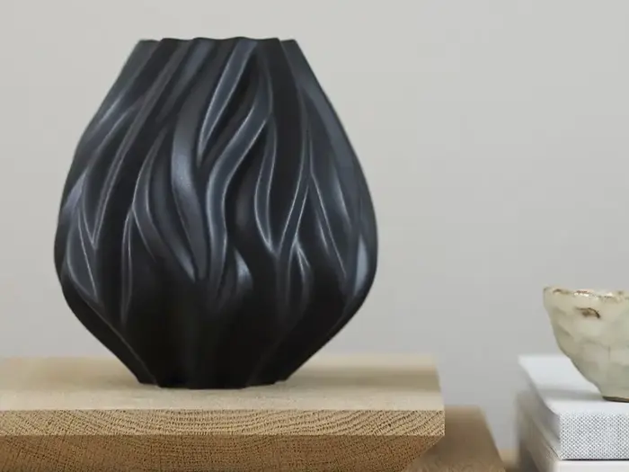 En sort, skulpturel vase med bølgende, flydende mønstre står på en træplade ved siden af en lille, lys keramikskål og stablede bøger.