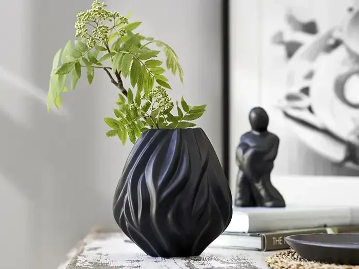 En struktureret sort vase med grønne bladgrene står på et rustikt bord med en lille sort skulptur og bøger sløret i baggrunden.
