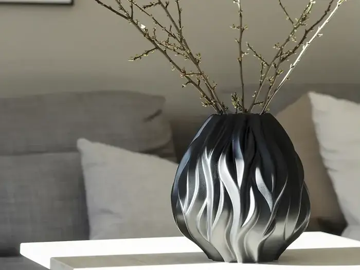 En sort og hvid skulpturel vase med bølgede mønstre indeholder et par nøgne grene, der står på et hvidt bord i en moderne stue med en grå sofa og hvide puder i baggrunden.