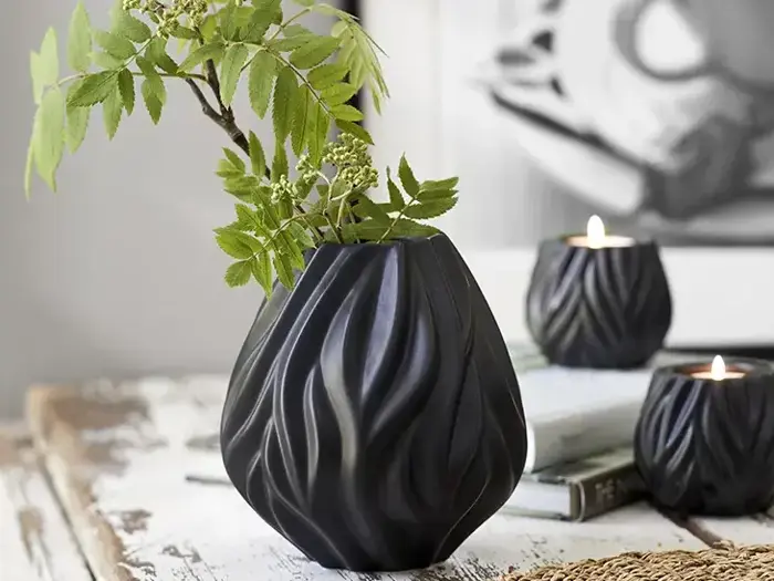 En sort, skulpturel vase med grønne bladgrene står på et rustikt hvidt bord ved siden af to matchende sorte lysestager med tændte stearinlys.