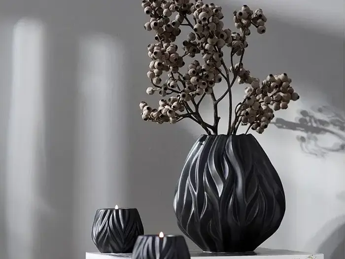 En sort vase med tørrede grene og to små tændte sorte stearinlys står på en hvid flade mod en lysegrå væg.