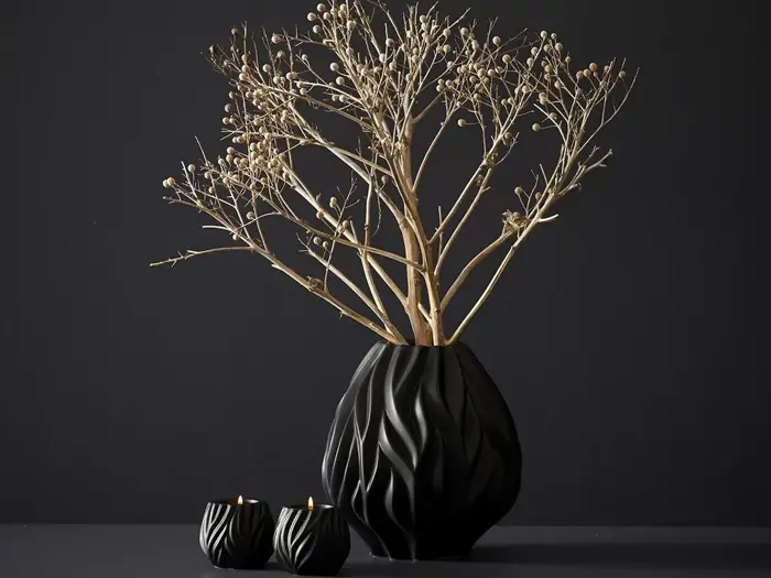 En sort vase med et bølget mønster indeholder tørre, bladløse grene. To små, matchende sorte lysestager med tændte stearinlys står ved siden af, alt sammen på en mørk baggrund.