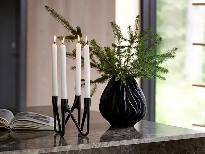 En sort zigzag-lysestage med fire tændte hvide lys står på en marmorplade ved siden af en sort vase med grønne fyrretræsgrene. En åben bog ligger i nærheden.