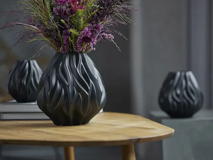 En sort vase med bølget tekstur og lilla og grønne blomster står på et træbord med to lignende tomme vaser i baggrunden.