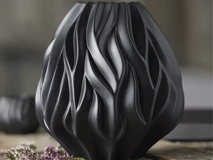 En matsort vase med flydende, skulpturelle bølgemønstre står på en træoverflade ved siden af små lyserøde blomster.