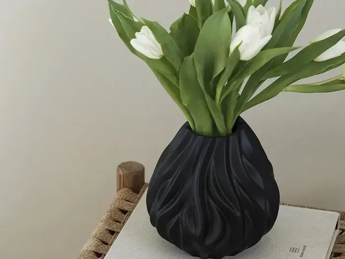 En sort skulpturel vase med hvide tulipaner og grønne blade står på en vævet skammel ved siden af en lys væg.