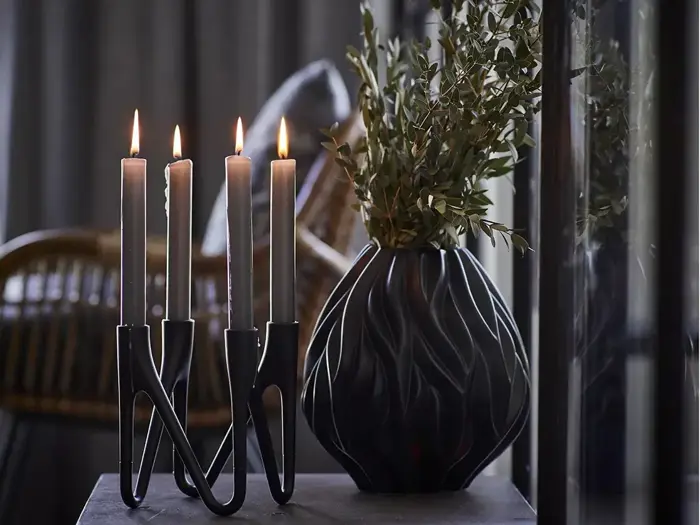 Fire tændte stearinlys i en moderne sort lysestage står ved siden af en mørk, struktureret vase med grønne blade på et bord med sløret baggrund.
