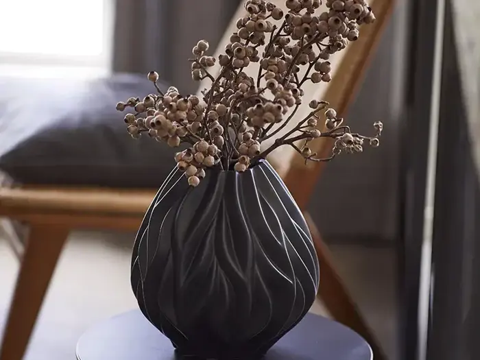 En sort, struktureret vase med dekorative, tørrede grene står på et mørkt, rundt bord i et svagt oplyst rum med en træstol i baggrunden.