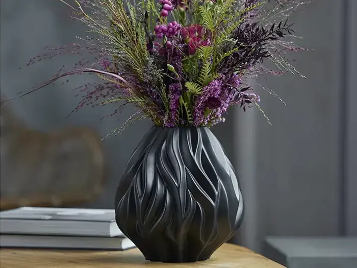 En sort, bølget vase fyldt med lilla og grønne kunstige blomster står på et træbord med bøger og en sløret baggrund bagved.