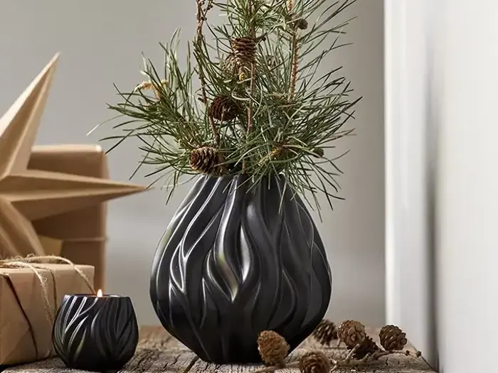En sort vase med et bølget mønster indeholder fyrretræsgrene og kogler på en rustik træoverflade ved siden af et lille lys, kogler og indpakkede gaver.