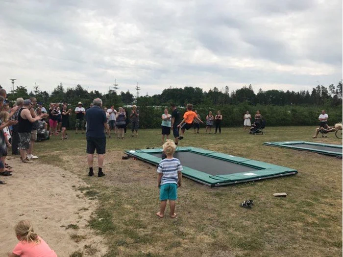 Folk samles udendørs omkring to trampoliner i jordhøjde, mens en ung dreng i forgrunden ser på. Det er overskyet, og omgivelserne ser ud til at være en park eller et åbent græsareal.
