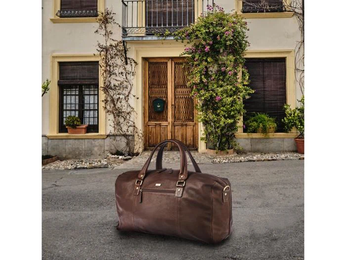 En stor brun lædertaske ligger på fortovet foran et hus med trædøre, potteplanter og klatrende slyngplanter.
