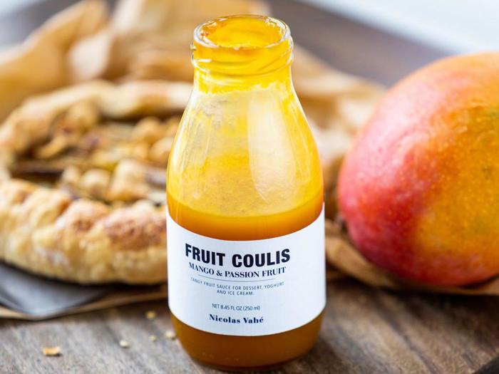 En flaske mango- og passionsfrugtcoulis står åben på en træoverflade, med en mango og et wienerbrød sløret i baggrunden.