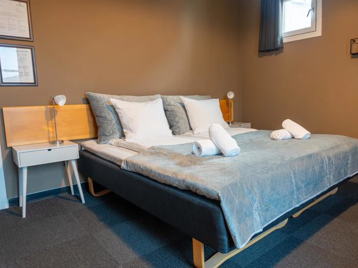 Moderne soveværelse med en pænt redt dobbeltseng, grå dyne, hvide puder og håndklæder, natborde med lamper og certifikater på den solbrune væg.