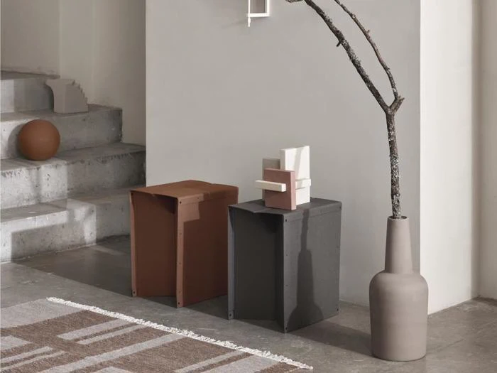 Minimalistisk interiør med betontrapper, to kantede skamler (brune og sorte), en abstrakt skulptur, en vase med en bar gren og et geometrisk tæppe på et poleret gulv.