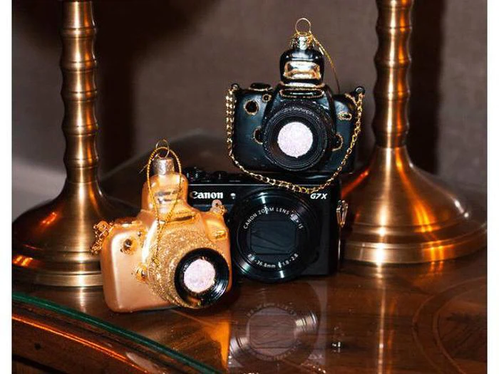To kameraformede ornamenter i guld og sort står ved siden af et ægte Canon-kamera på et træbord med gyldne lampefødder i baggrunden.