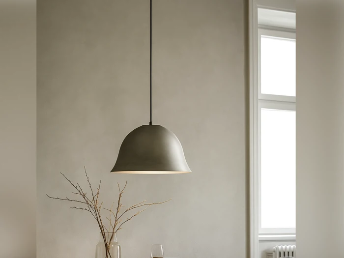 En minimalistisk pendellampe hænger fra loftet ved siden af et vindue med tørrede grene i en vase på bordet nedenfor mod en neutral væg.