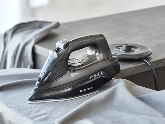 Et sort Nordic Sense-dampstrygejern står på et gråt strygebræt med en matchende grå skjorte ved siden af sin opladningsbase på en bordplade.