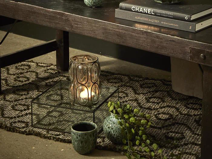 Et stearinlys i en glasholder står på en glasbakke under et mørkt bord sammen med en lille grøn plante og en keramikkop, og bøgerne er stablet oven på bordet.