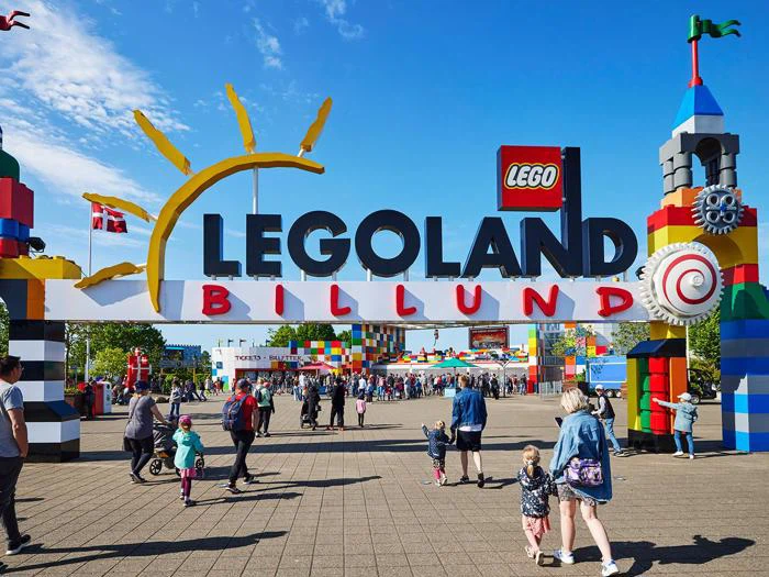 Indgang til LEGOLAND Billund med et stort, farverigt skilt, dekorationer med LEGO-tema og besøgende, der går mod parken under en lyseblå himmel.