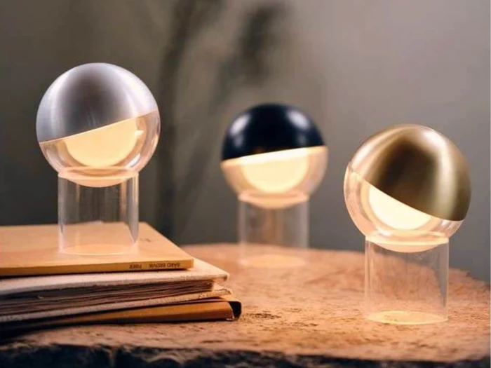 Tre moderne, kugleformede lamper med matte skærme og metalliske halvskaller, der udstråler varmt lys, står på en stak bøger og en træoverflade i en hyggelig omgivelse.