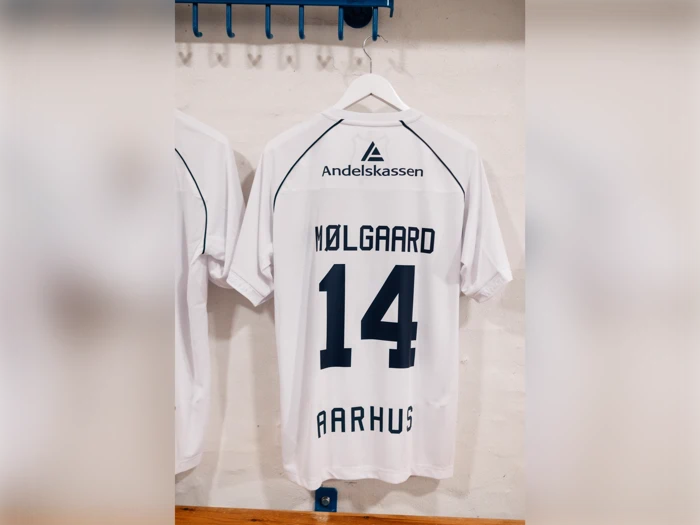 Hvid fodboldtrøje med NØLGAARD og 14 på ryggen samt Andelskassen-logoet og AARHUS trykt på, hængende på et vægstativ.