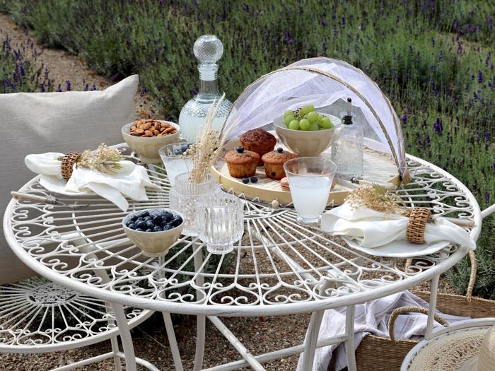 Et hvidt metalbord dækket udendørs med muffins, vindruer, nødder, blåbær, vand og mælk. Tørrede blomster pynter på servietterne, og over frugt og muffins ligger et låg. Lavendel blomstrer i baggrunden