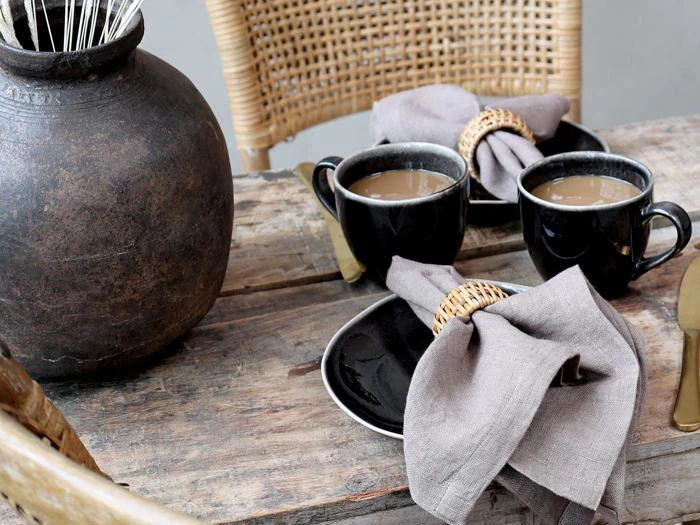 To sorte kopper kaffe med mælk på underkopper, hver med en foldet linnedserviet og flettet ring, sat på et rustikt træbord med en stor mørk vase og flettede stole.