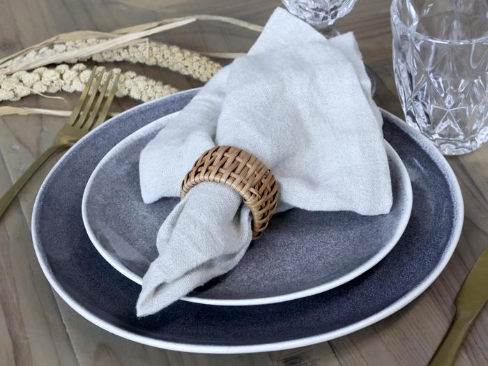 En borddækning med to stablede blågrå tallerkener, en beige hørserviet i en vævet ring, guldbestik, krystalglas og dekorative tørrede planter.