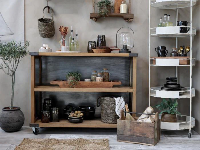 Et rustikt køkken med træhylder med krukker, skåle, kurve, redskaber og planter sammen med sorte tallerkener på et hvidt metalstativ og en trækasse med brød og grønt.