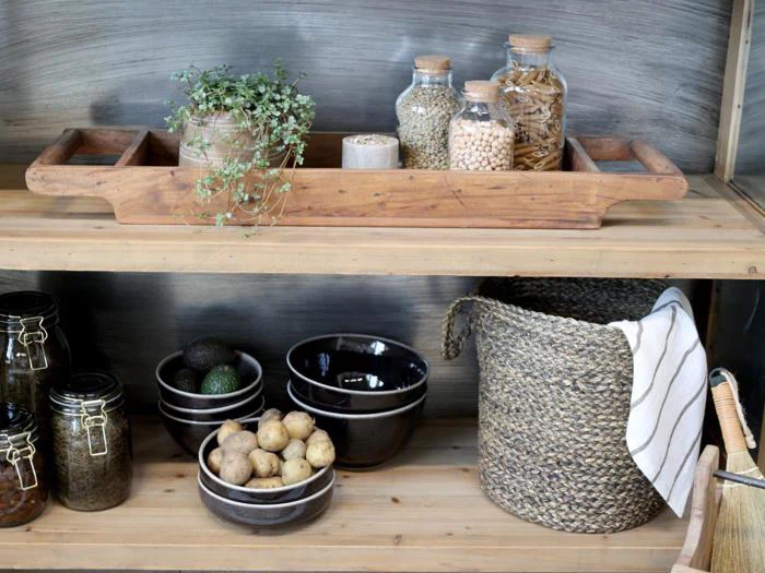 På træhylder står der krukker med korn, en potteplante, sorte skåle med kartofler og avocadoer, glaskrukker og en flettet kurv med et stribet håndklæde.