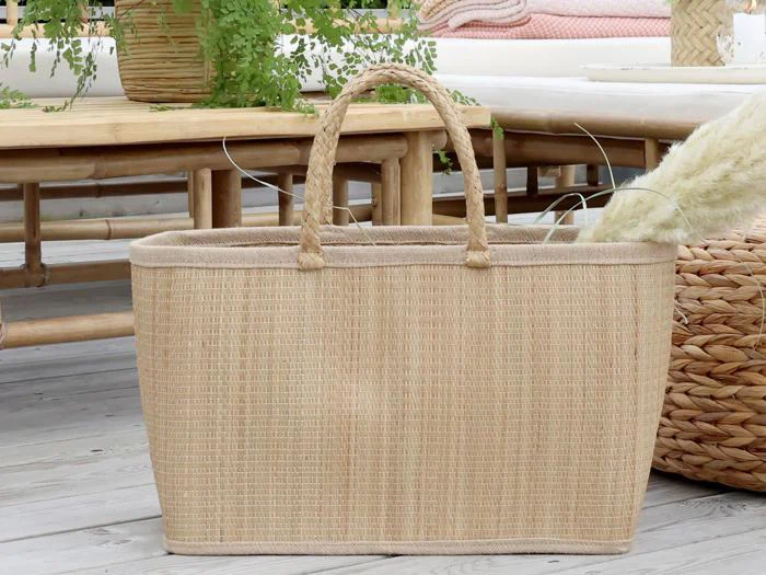 En vævet beige tote bag med håndtag ligger på et trægulv nær kurvemøbler og grønne planter i et hyggeligt, lyst rum.