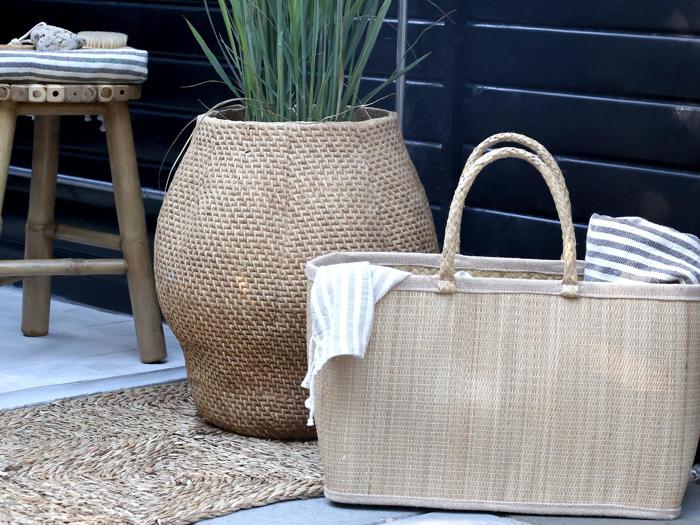 En flettet kurv med håndtag og stribede klude står på et vævet tæppe ved siden af en stor krukke med højt græs og en lille træskammel.