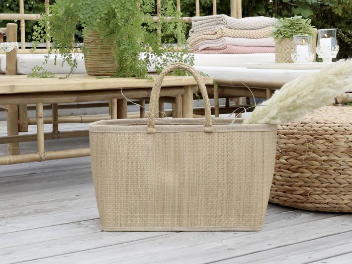 En flettet kurv med håndtag står på en træterrasse omgivet af havemøbler, grønne planter og sammenfoldede tæpper.