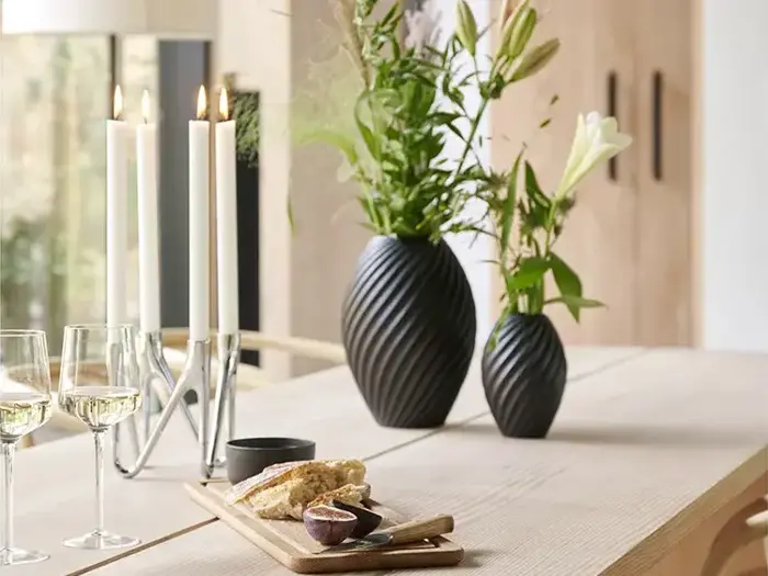 Et lyst træbord med vinglas, en lysestage med tændte stearinlys, et skærebræt med brød og redskaber og to sorte vaser med grønne og hvide blomster.