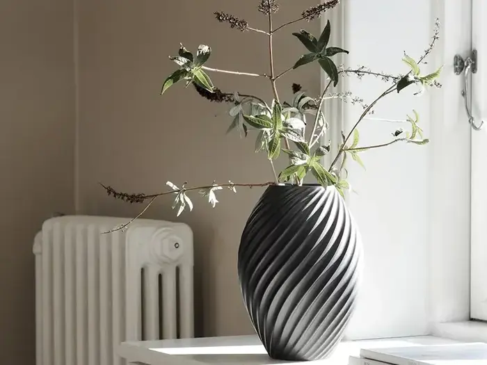 En sort, struktureret vase med grønne bladgrene står på et hvidt bord ved et solbeskinnet vindue med en radiator i baggrunden.