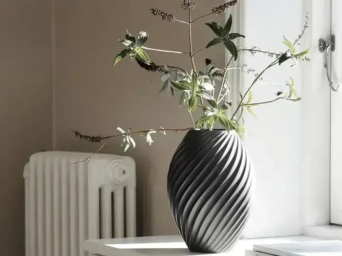 En mørkegrå, spiralformet vase med grene med blade står på en hvid vindueskarm ved siden af en radiator.