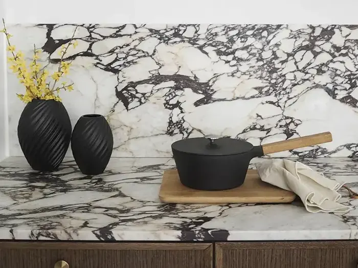 En bordplade af marmor med sorte årer, to sorte vaser med gule blomster, en tildækket sort gryde med træhåndtag på et bræt og et sammenfoldet klæde ved siden af.