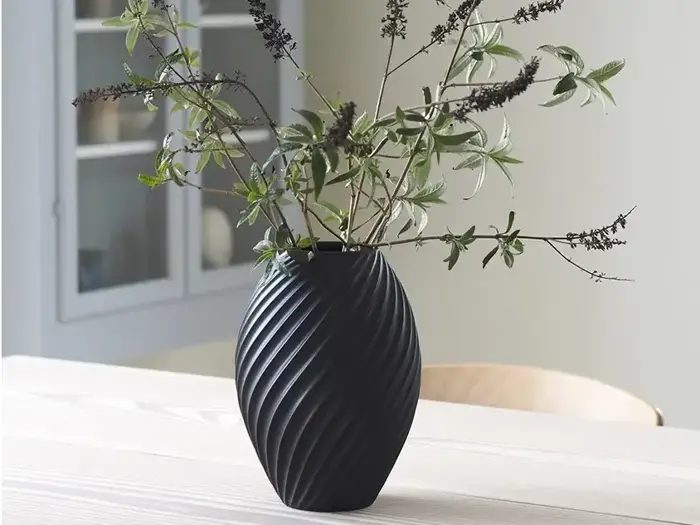 En sort, struktureret vase med diagonale riller rummer sparsomme grønne og brune bladgrene, der er placeret på et lyst træbord indendørs.