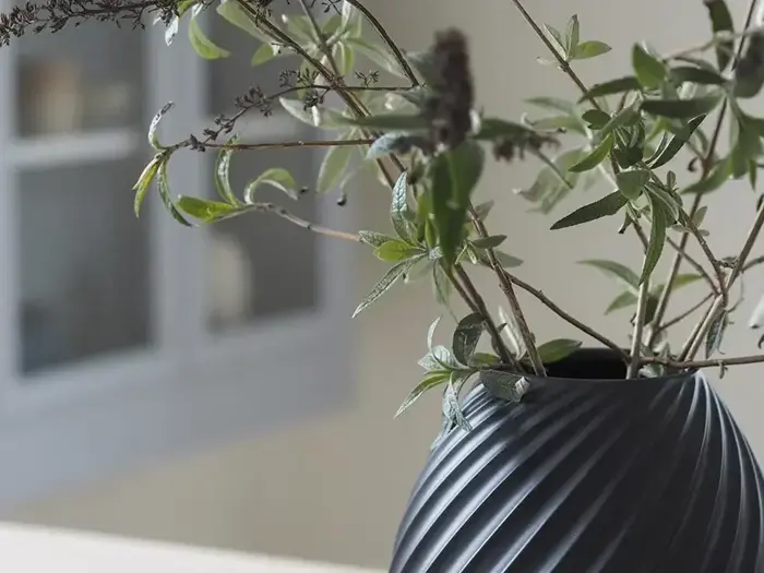 Et nærbillede af grønne bladgrene i en mørk, riflet vase med en sløret baggrund, der viser en del af et vindue eller et skab.