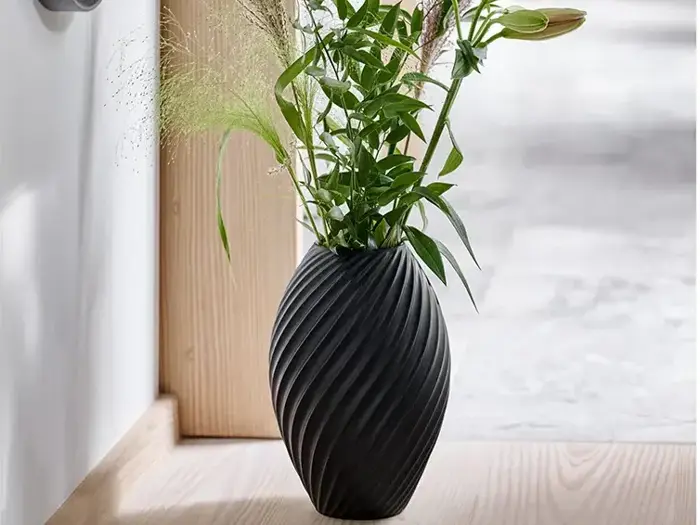 En sort, struktureret vase med spiralmønster rummer grønne bladplanter og græsser, placeret på et lyst trægulv nær et vindue.