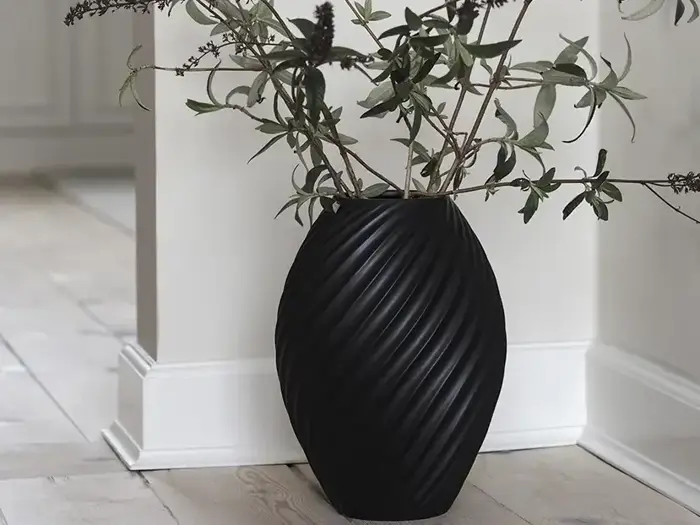 En sort, struktureret vase med grønne grene og mørke knopper står på et lyst trægulv ved siden af en hvid væg og fodliste.
