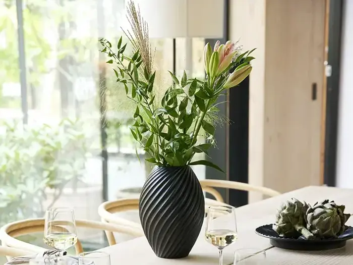 En sort, struktureret vase med grønne blade og lyserøde liljer på et spisebord, omgivet af vinglas og en tallerken med artiskokker.