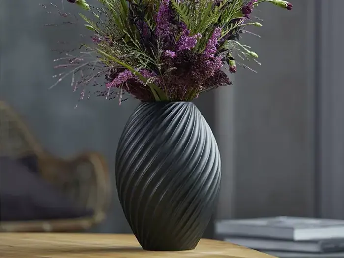 En sort, struktureret vase med diagonale riller holder en buket lilla blomster og grønt, placeret på en træoverflade indendørs.