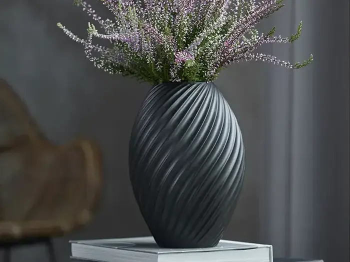 En sort, struktureret vase med et spiraldesign med lilla lyngblomster står oven på en lukket bog.